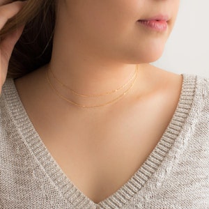 Puede incluir: Dos delicados collares dorados en capas. Los collares son finos y cuelgan alrededor del cuello. La imagen muestra un primer plano de los collares sobre una persona que lleva un su&eacute;ter gris.