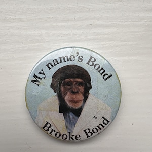 May include: A circular badge with a light blue backdrop, displaying a chimpanzee in a white jacket and black bow tie. The text "My name's Bond" and "Brooke Bond" are printed around the chimpanzee image.
