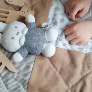 Op de afbeelding: Een bruine en witte patchwork baby speelmat met een grijs en wit knuffelkonijn. De speelmat heeft verschillende texturen en patronen, waaronder sterren, strepen en effen kleuren.