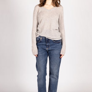 May include: A woman wearing a light gray v-neck sweater and blue denim jeans. She is wearing white sneakers.
