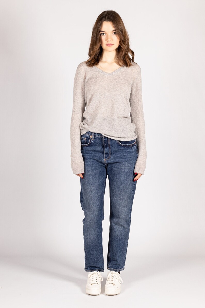 May include: A woman wearing a light gray v-neck sweater and blue denim jeans. She is wearing white sneakers.