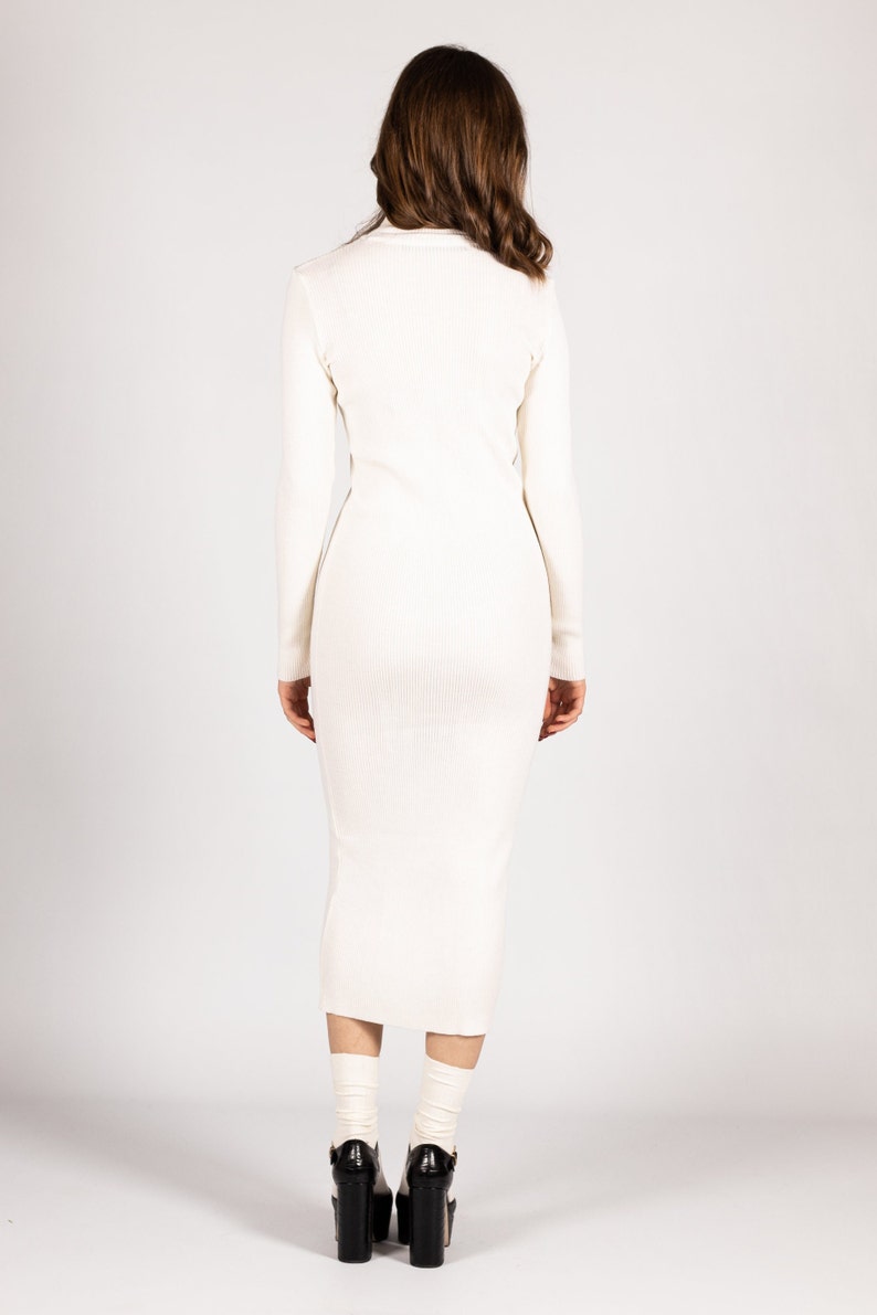 K&ouml;nnte beinhalten: Ein wei&szlig;es geripptes Midikleid mit langen &Auml;rmeln. Das Kleid ist figurbetont und hat einen leicht ausgestellten Rock. Das Model tr&auml;gt das Kleid mit schwarzen Plateau-Heels.