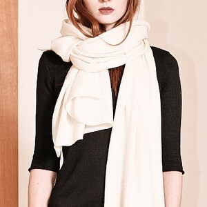 May include: A white knitted scarf draped around a person's neck. The scarf is long and flowing, with a soft, drapey texture. The person is wearing a black top and has long, brown hair.