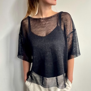 Peut inclure: Un haut noir transparent à manches courtes et à coupe ample. Le haut est fait d'une matière semblable à de la résille, révélant un caraco noir en dessous. L'encolure est large et arrondie. Le modèle porte un pantalon blanc.