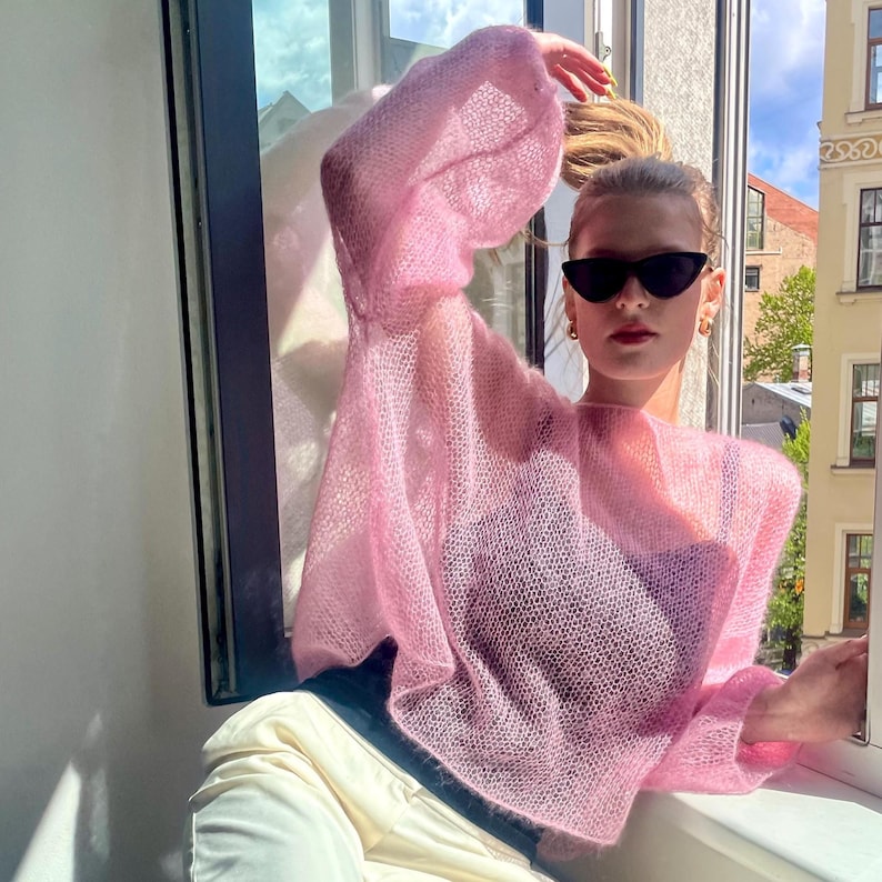 Puede incluir: Un su&eacute;ter rosa claro y transparente con mangas largas y voluminosas. La persona lleva gafas de sol negras y pendientes de aro dorados. El su&eacute;ter se lleva con un top negro y pantalones color crema. El su&eacute;ter tiene un dise&ntilde;o de punto abierto.