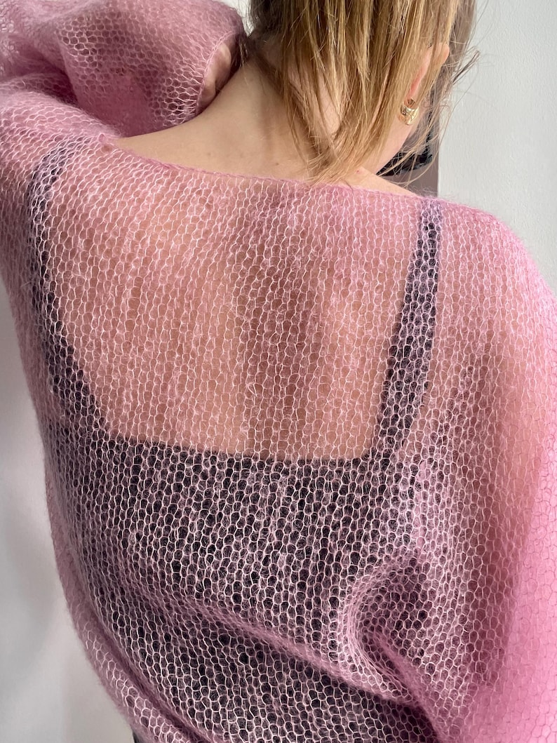 Puede incluir: Un su&eacute;ter rosa claro de tejido abierto. El su&eacute;ter est&aacute; hecho de un material transparente y delicado, con un ajuste holgado y aireado. El su&eacute;ter se usa sobre una parte superior negra, creando un contraste de color y textura. El aspecto general es suave y femenino.