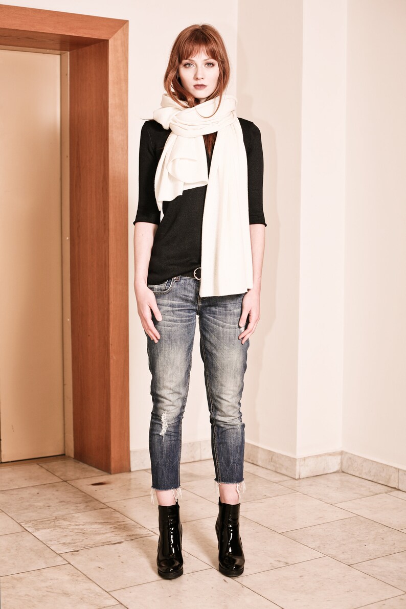 May include: A woman wearing a black long-sleeved shirt, white scarf, blue jeans, and black ankle boots.