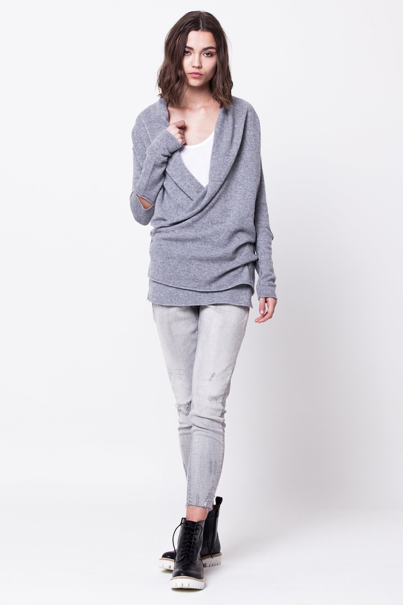 May include: A gray wrap sweater with long sleeves and a cowl neck. The sweater has a distressed look with frayed edges. The model is wearing the sweater with light gray jeans and black ankle boots.