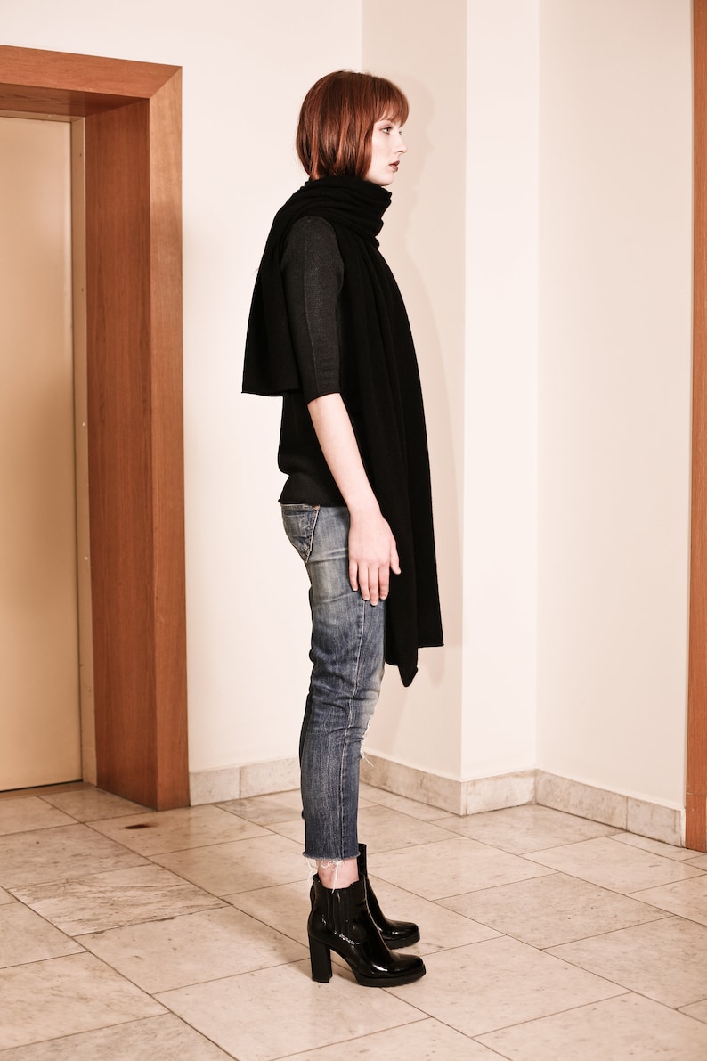 May include: A woman wearing a black mock neck shirt and blue jeans with a black scarf. She is wearing black heeled boots.