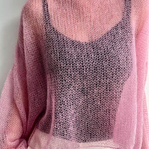 Puede incluir: Un su&eacute;ter de punto rosa claro y transparente con cuello ancho y mangas largas. El su&eacute;ter se usa sobre una camiseta negra y pantalones blancos. El tejido tiene un patr&oacute;n de panal.