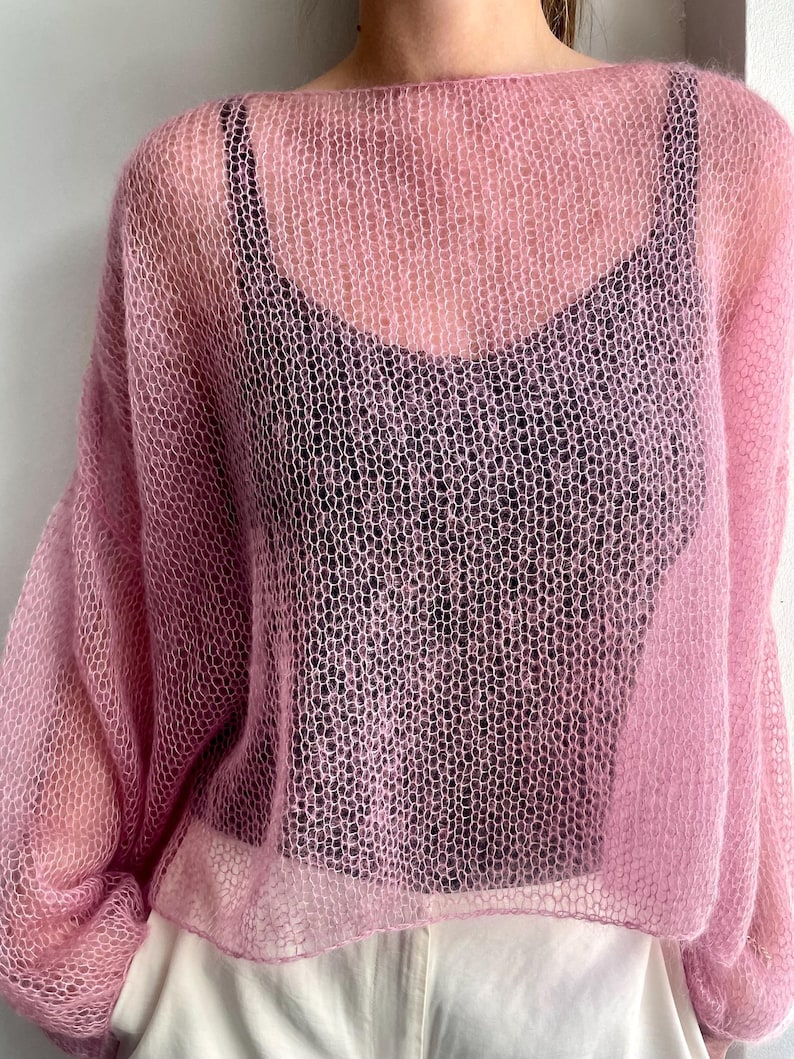 Puede incluir: Un su&eacute;ter de punto rosa claro y transparente con cuello ancho y mangas largas. El su&eacute;ter se usa sobre una camiseta negra y pantalones blancos. El tejido tiene un patr&oacute;n de panal.