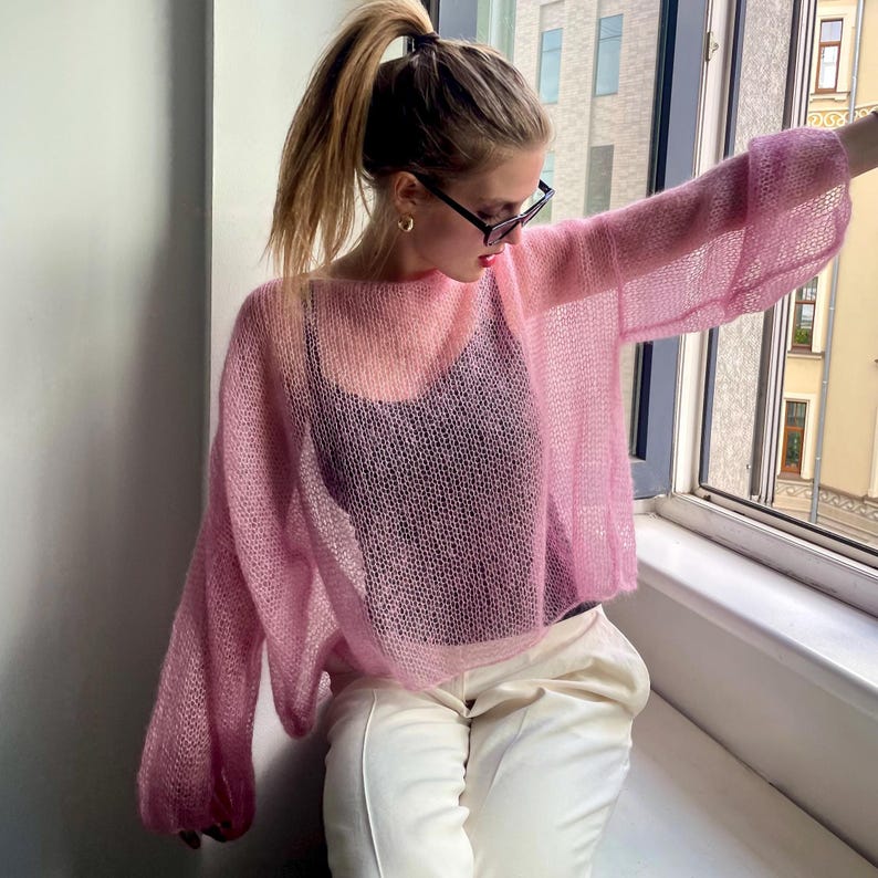 Puede incluir: Un su&eacute;ter de punto rosa claro y transparente con mangas largas y acampanadas. El su&eacute;ter se usa sobre una camiseta sin mangas negra y se combina con pantalones blancos. El su&eacute;ter tiene un ajuste holgado y de gran tama&ntilde;o.