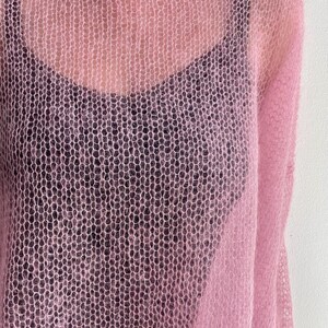 Puede incluir: Un su&eacute;ter rosa claro de tejido abierto con cuello redondo. El su&eacute;ter est&aacute; superpuesto a una camiseta sin mangas negra. La tela de punto tiene una apariencia delicada y aireada. Las mangas del su&eacute;ter son visibles en el lado derecho de la imagen.
