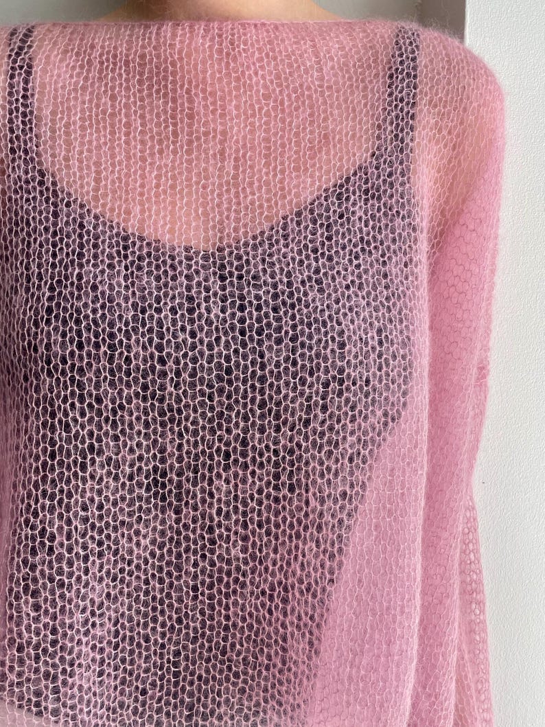 Puede incluir: Un su&eacute;ter rosa claro de tejido abierto con cuello redondo. El su&eacute;ter est&aacute; superpuesto a una camiseta sin mangas negra. La tela de punto tiene una apariencia delicada y aireada. Las mangas del su&eacute;ter son visibles en el lado derecho de la imagen.