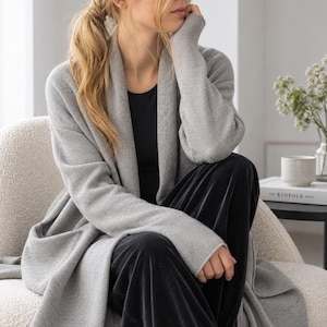May include: A long-sleeved light gray open-front cardigan with a wide shawl collar, worn over a plain black top and black velvet trousers. The knit garment features a smooth texture, long sleeves with ribbed cuffs, and a relaxed, draped silhouette.