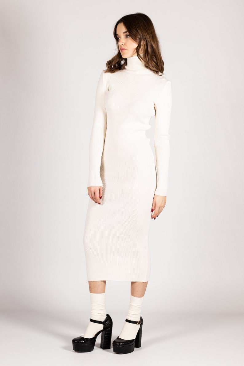 K&ouml;nnte beinhalten: Ein wei&szlig;es, geripptes Rollkragenpulloverkleid mit langen &Auml;rmeln. Das Kleid ist figurbetont und reicht bis zur Mitte der Wade. Das Model tr&auml;gt schwarze Plateauschuhe mit wei&szlig;en Socken.