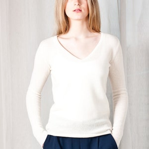 May include: A woman wearing a white v-neck sweater and dark blue pants. She is standing in front of a white background.