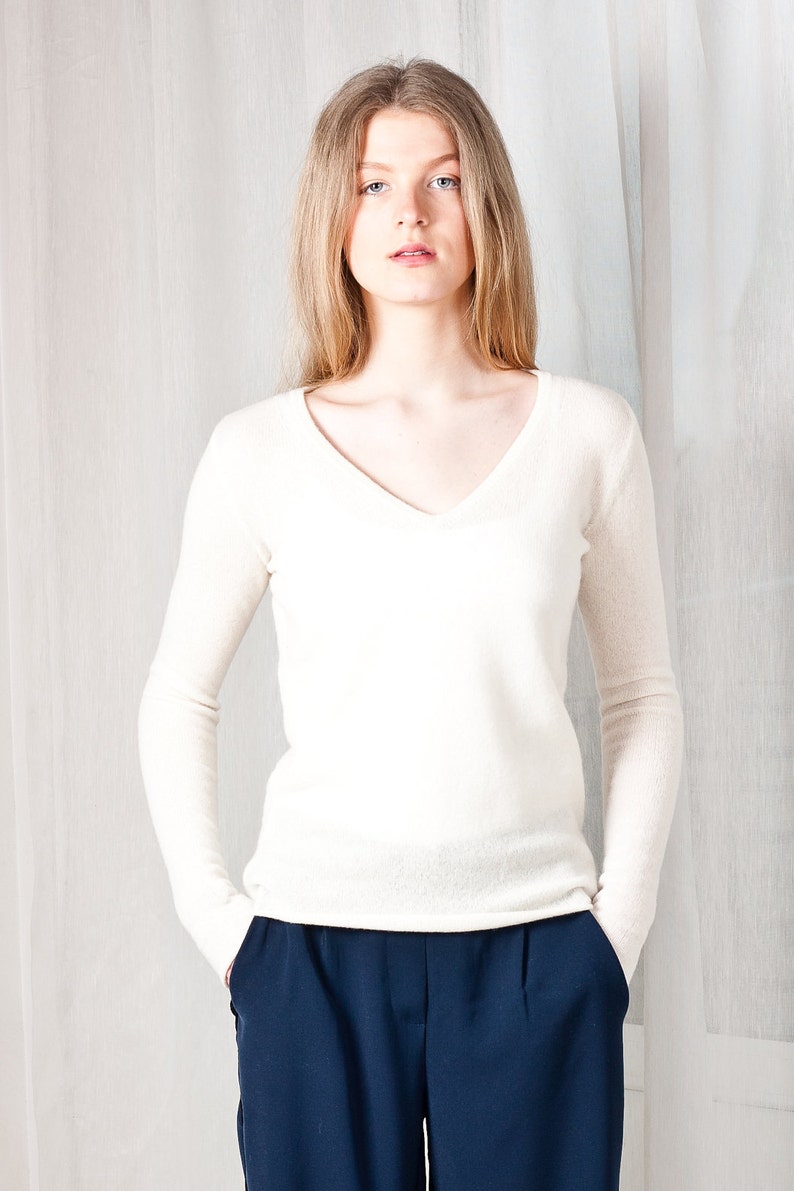 May include: A woman wearing a white v-neck sweater and dark blue pants. She is standing in front of a white background.