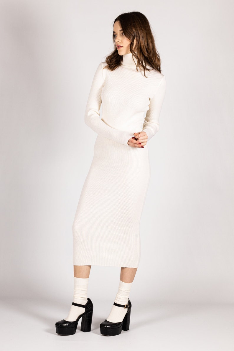 K&ouml;nnte beinhalten: Ein wei&szlig;es Rollkragenpulloverkleid mit gerippter Struktur. Das Kleid ist figurbetont und reicht bis zur Mitte der Wade. Das Model tr&auml;gt schwarze Plateau-Heels mit wei&szlig;en Socken.