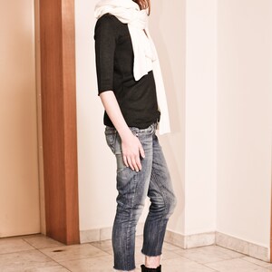 May include: A woman wearing a black long-sleeved shirt, white scarf, blue jeans, and black heeled ankle boots.
