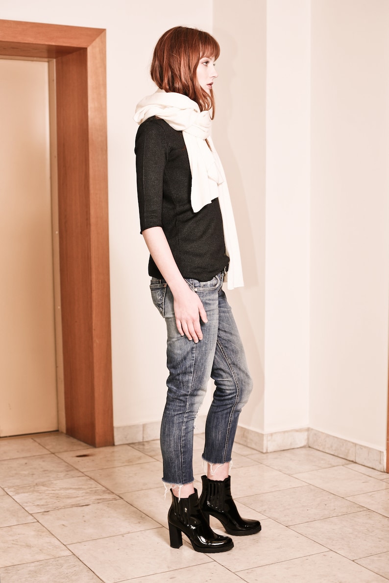 May include: A woman wearing a black long-sleeved shirt, white scarf, blue jeans, and black heeled ankle boots.