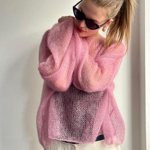 Puede incluir: Un su&eacute;ter de punto rosa claro y transparente con mangas largas y voluminosas. El su&eacute;ter tiene un tejido delicado y abierto, creando una apariencia suave y aireada. La modelo lleva gafas de sol y tiene el pelo recogido.