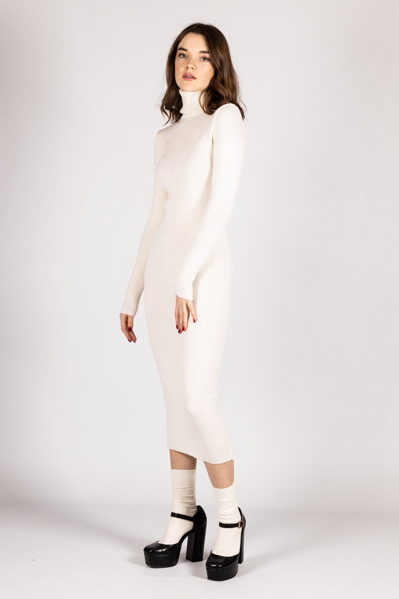 K&ouml;nnte beinhalten: Ein wei&szlig;es, geripptes Rollkragenpulloverkleid mit langen &Auml;rmeln. Das Kleid ist figurbetont und reicht bis zur Mitte der Wade. Das Model tr&auml;gt wei&szlig;e Socken und schwarze Plateauschuhe.