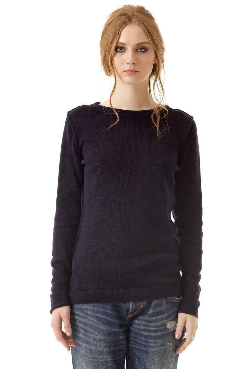 May include: A long-sleeved, dark navy blue knit sweater featuring a wide boat neckline and visible seam detailing along the shoulders and sides. The sweater is paired with blue denim jeans that have a distressed, worn-in texture and a faded wash.