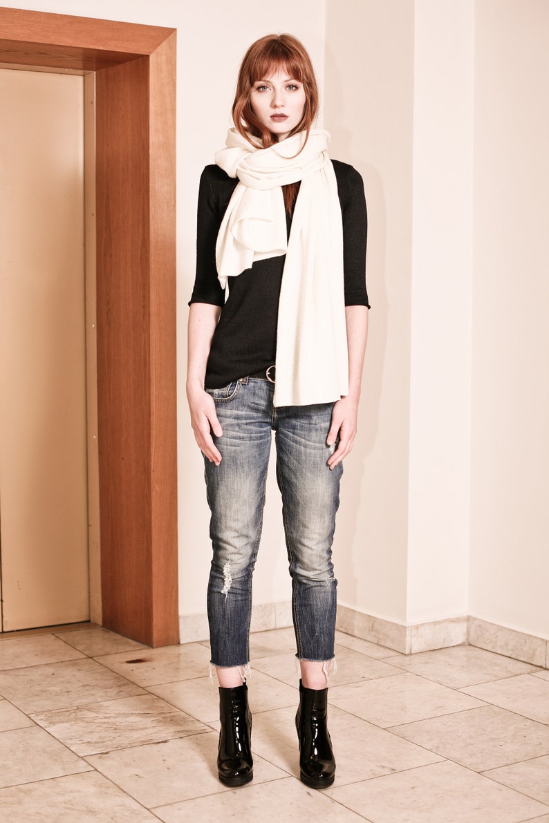 May include: A woman wearing a white scarf, a black long-sleeved shirt, blue jeans, and black ankle boots.