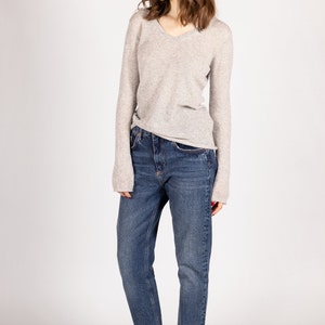 May include: A woman wearing a light gray v-neck sweater and blue jeans. She is wearing white sneakers.