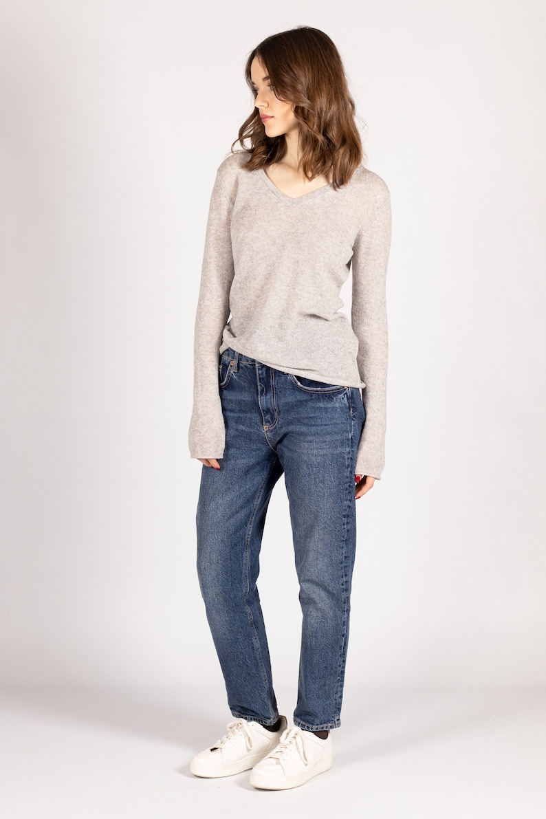 May include: A woman wearing a light gray v-neck sweater and blue jeans. She is wearing white sneakers.