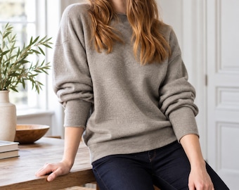 May include: A long-sleeved, crew-neck sweater in a solid light taupe color, featuring a relaxed fit with dropped shoulders and ribbed cuffs. The garment is paired with dark blue denim jeans, creating a simple and neutral ensemble.