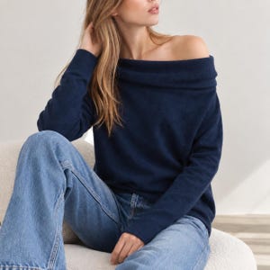 May include: A dark navy blue long-sleeve sweater featuring an off-the-shoulder neckline with a wide folded collar design, paired with light blue denim jeans. The sweater has a soft texture and long sleeves that extend to the wrists.