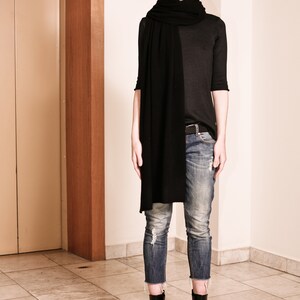 May include: A woman wearing a black turtleneck sweater, a black scarf, blue jeans, and black ankle boots. She is standing in a hallway with a wooden door frame.