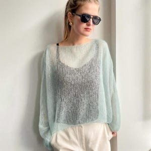 Könnte beinhalten: Ein hellblaues, durchsichtiges Strickoberteil mit weitem Ausschnitt. Der Pullover wird über einem schwarzen Top getragen und mit cremefarbenen Hosen kombiniert. Das Model trägt eine schwarze Sonnenbrille und goldene Ohrringe.