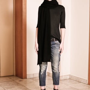 May include: A woman wearing a black long-sleeved shirt, black scarf, blue jeans, and black heeled boots. She is standing in front of a wooden door.