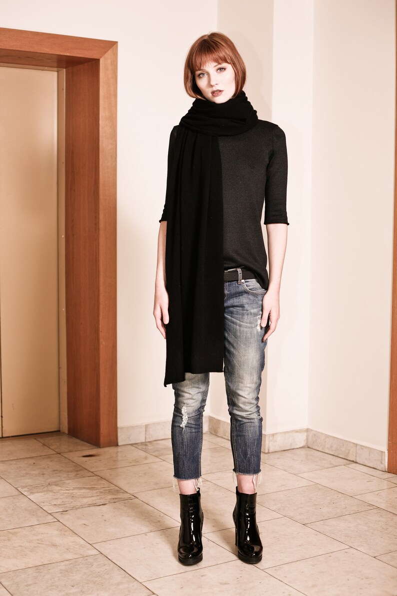 May include: A woman wearing a black long-sleeved shirt, black scarf, blue jeans, and black heeled boots. She is standing in front of a wooden door.