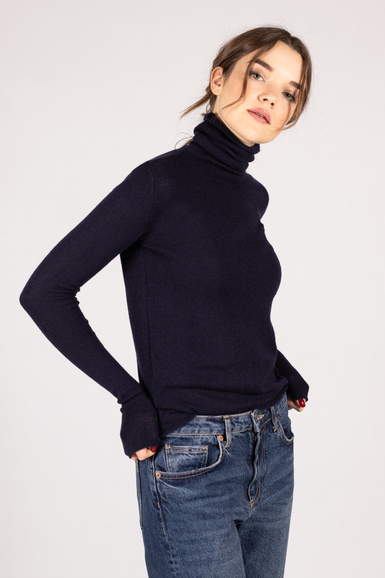 May include: A dark navy blue long-sleeve turtleneck sweater with a fitted silhouette and ribbed texture, paired with classic blue denim jeans featuring a button fly and traditional five-pocket construction with copper-colored metal rivets.