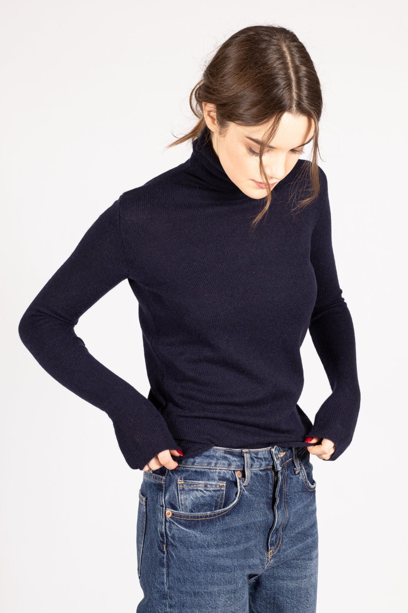 May include: A dark navy blue long-sleeve turtleneck sweater with a fine knit texture, paired with classic blue denim jeans featuring a button fly and metal rivets. The sweater has a fitted silhouette and long sleeves that extend past the wrists.