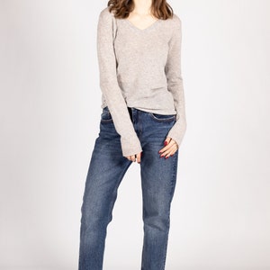 May include: A woman wearing a light gray long-sleeved sweater and blue jeans. She is standing with her arms at her sides and looking at the camera.