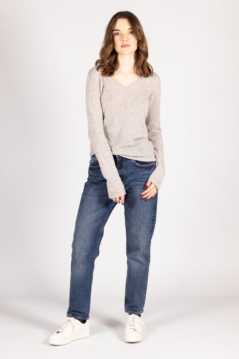 May include: A woman wearing a light gray long-sleeved sweater and blue jeans. She is standing with her arms at her sides and looking at the camera.
