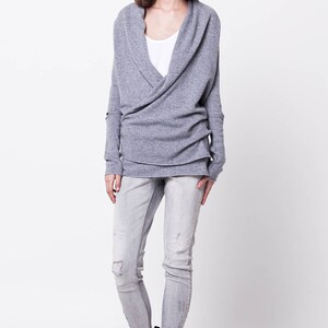 May include: A gray wrap sweater with long sleeves and a cowl neck. The sweater is worn over a white t-shirt and paired with light blue jeans and black lace-up boots.