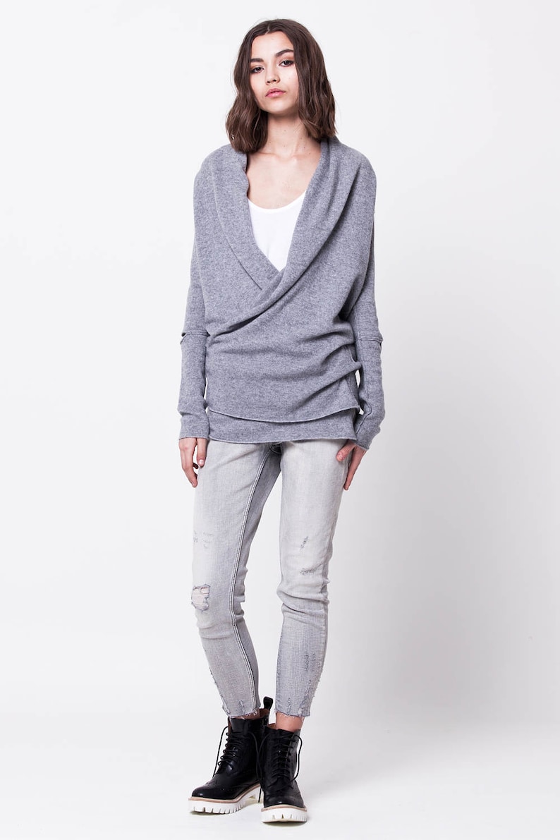 May include: A gray wrap sweater with long sleeves and a cowl neck. The sweater is worn over a white t-shirt and paired with light blue jeans and black lace-up boots.