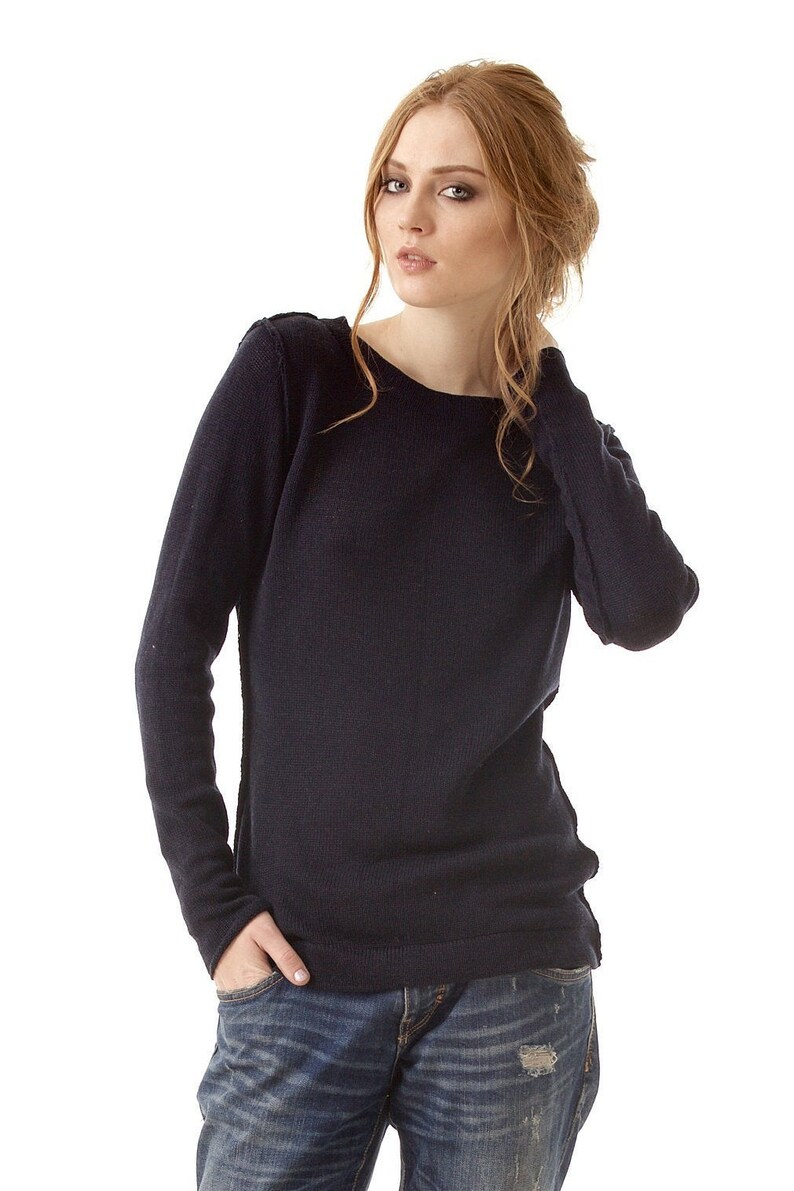 May include: A long-sleeved navy blue knit sweater featuring a wide boat neckline and visible seam detailing along the shoulders and sides, paired with distressed blue denim jeans that have a worn-in texture and a small rip on the front thigh area.