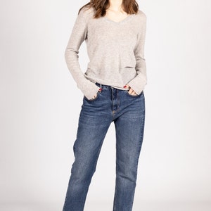 May include: A woman wearing a light gray v-neck sweater and blue jeans. She is standing with her hands in her pockets and looking at the camera.
