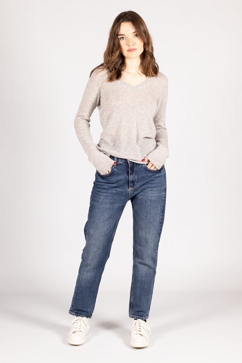 May include: A woman wearing a light gray v-neck sweater and blue jeans. She is standing with her hands in her pockets and looking at the camera.