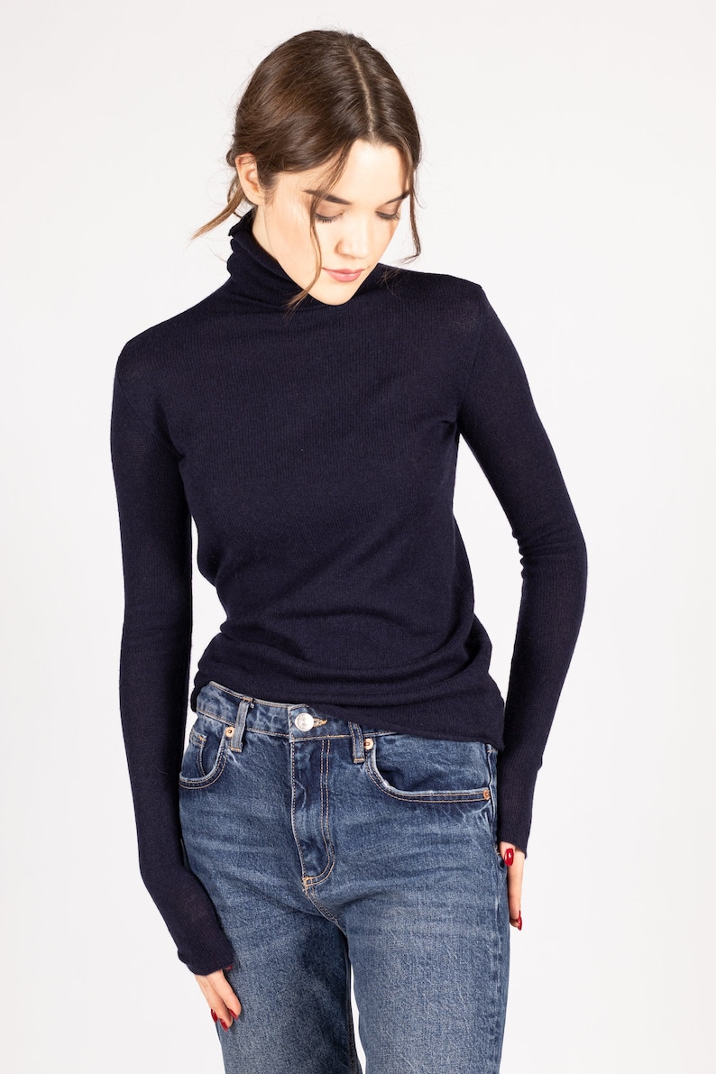 May include: A dark navy blue long-sleeve turtleneck sweater with a ribbed knit texture and extended cuffs that cover the wrists, paired with dark blue denim jeans featuring a metal button closure and classic five-pocket styling.
