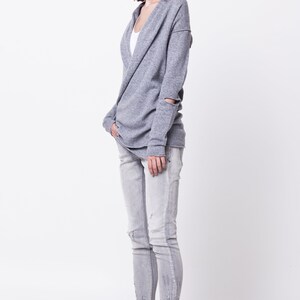 May include: A woman wearing a gray knit cardigan with a cowl neck and ripped jeans. She is wearing black ankle boots with white soles.