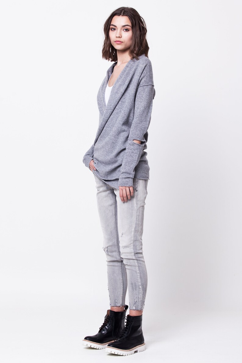 May include: A woman wearing a gray knit cardigan with a cowl neck and ripped jeans. She is wearing black ankle boots with white soles.