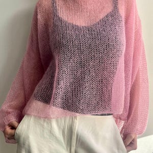 Puede incluir: Un su&eacute;ter de punto rosa claro y transparente con cuello ancho y mangas largas. El su&eacute;ter se usa sobre una camiseta negra y se combina con pantalones blancos. El tejido tiene un tejido delicado y abierto.